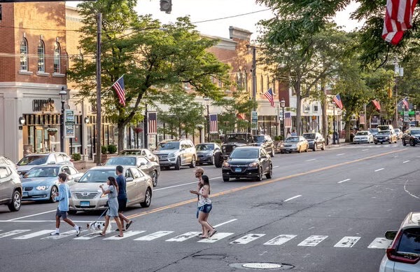 Pedestrians crossing a shopping street lined with parked cars